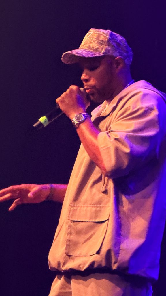 A person wearing a cap and jacket performing on stage under colorful lighting.