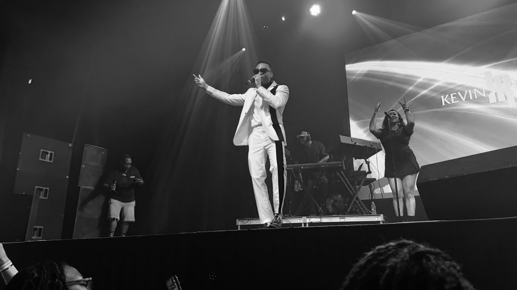 A performer on stage in a white suit gesturing with his arms, with a large "KEVIN" projection behind him and other performers visible.