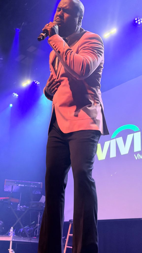 A man in a pink suit performing on stage, with the "VIVI" logo visible behind him.