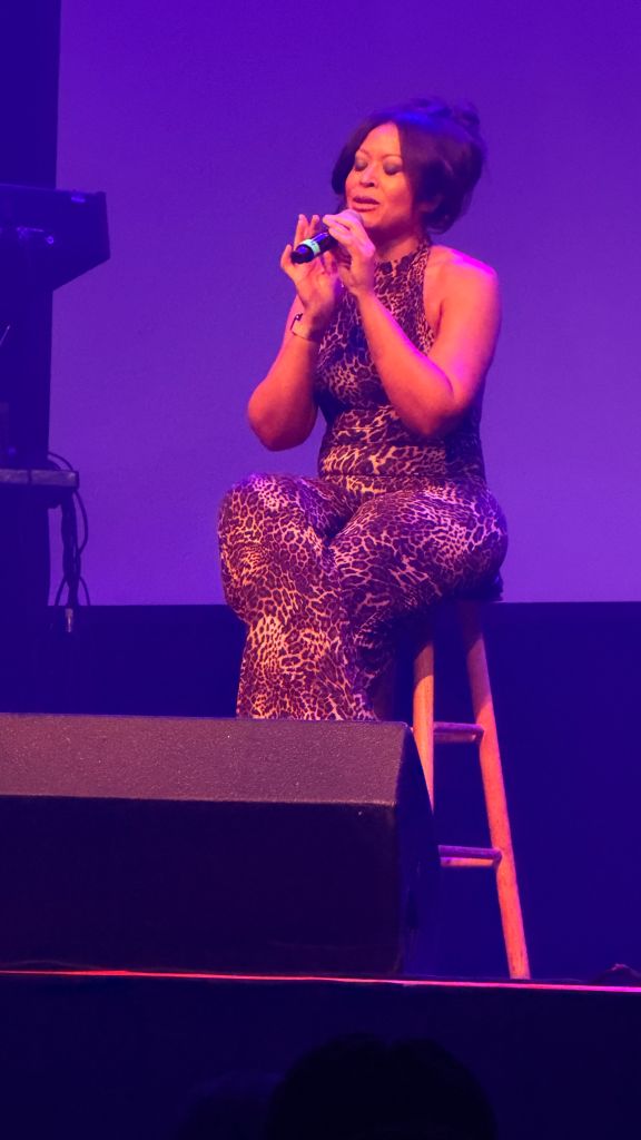 A woman in a leopard print outfit singing into a microphone on stage under purple lighting.