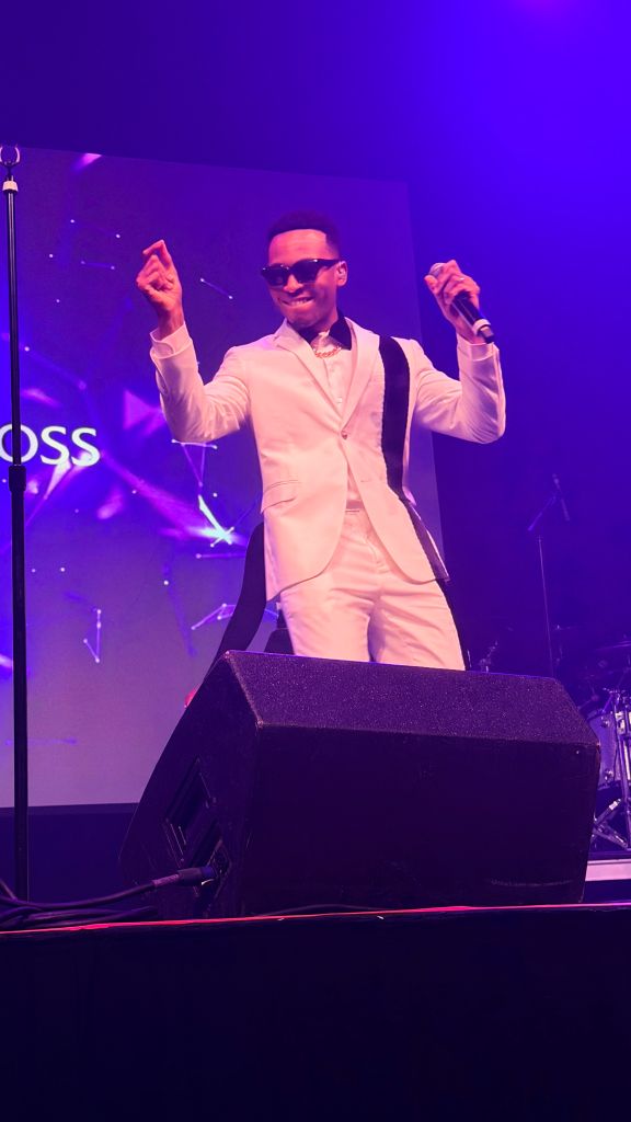 A man in a white suit and sunglasses performing on stage with a microphone and speaker behind him.