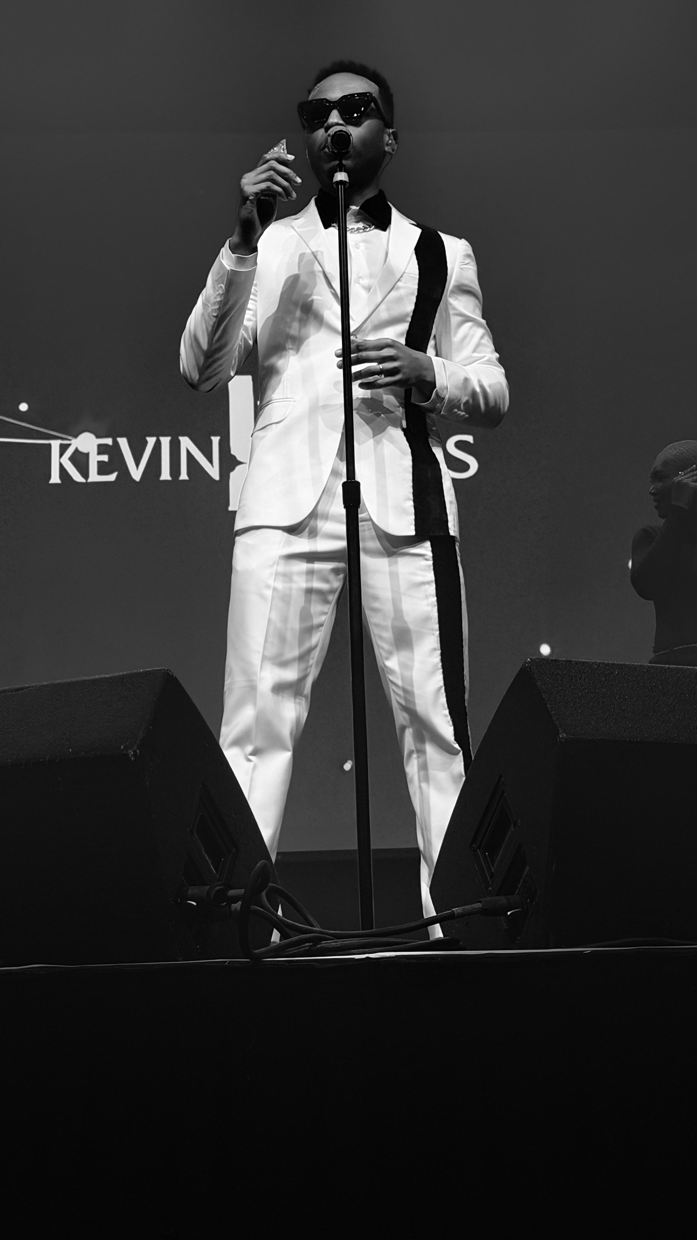 A person wearing a white suit and sunglasses performing on stage, with the text "KEVIN JONAS" visible behind them.