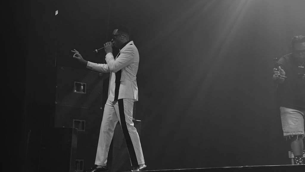 A person in a white suit performing on a stage in a dark setting, holding a microphone and gesturing with their hands.